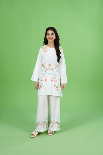 A woman standing against a green background, wearing a white crop shirt with embroidery details and matching white straight pants.