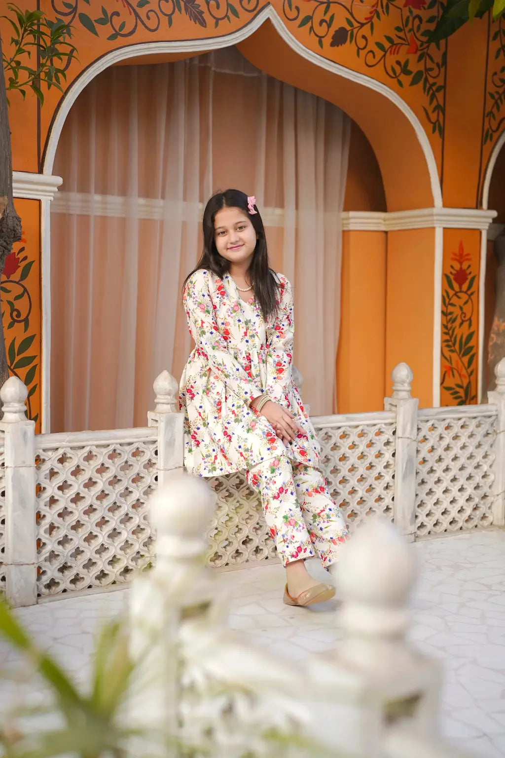 Young girl in a floral dress sitting on a decorative bench with an ornate orange and white background.