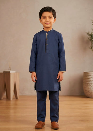 Young boy wearing a navy kurta with brown shoes in a room with wooden floor and beige wall.