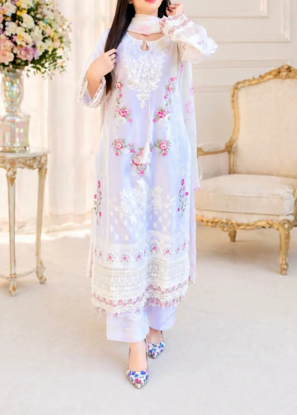 A woman posing in a 3-piece white embroidered cotton lawn suit. The outfit features pink floral embroidery on the shirt, organza cutwork sleeves, and a heavy lace border on the daman. Paired with straight-cut trousers and a printed floral dupatta. Perfect for summer fashion and festive wear.