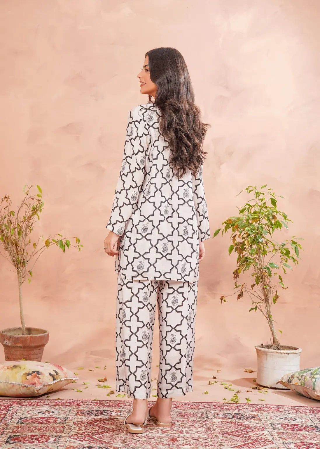 Woman in a patterned outfit standing in a room with a pink wall and decorative plants.