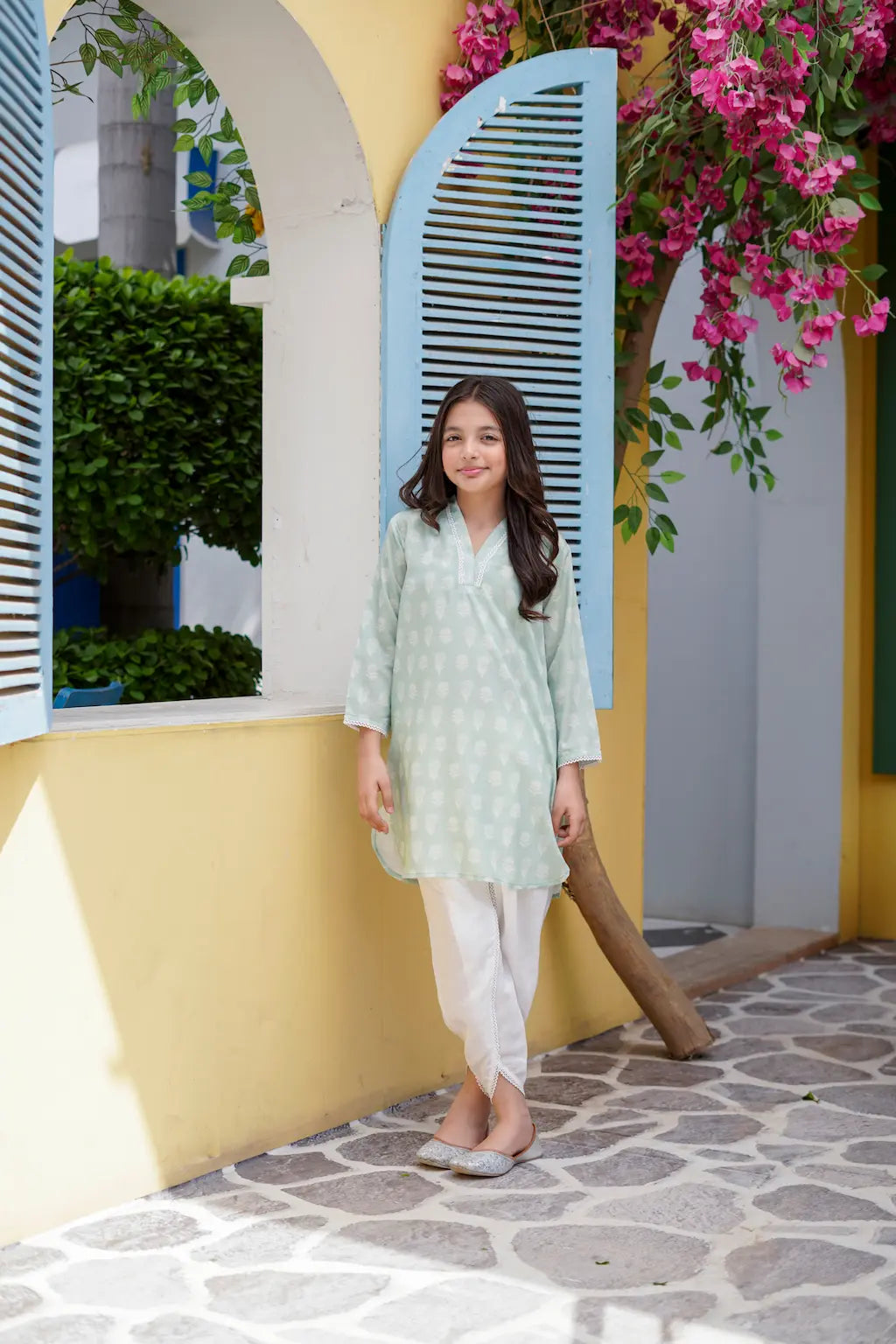 Young girl in a light green traditional outfit standing in front of a yellow wall with pink flowers.