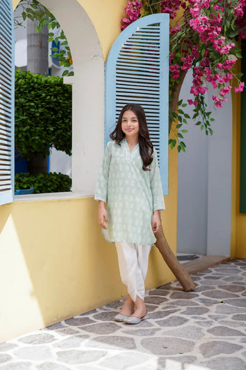 Young girl in a light green traditional outfit standing in front of a yellow wall with pink flowers.