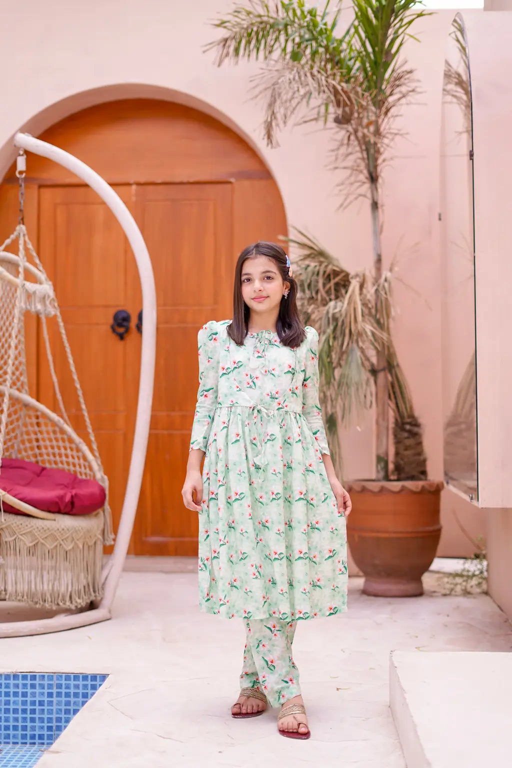 A young girl in a floral dress standing in front of a poolside house with a swing and palm tree.