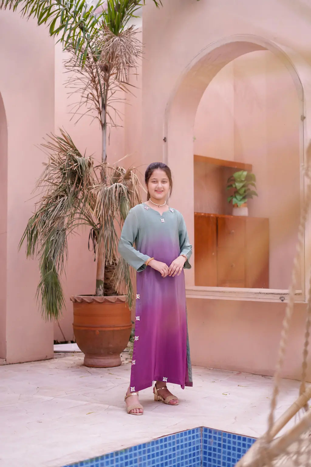 Girl in a gradient dress standing by a pool with a pink wall and plant in the background