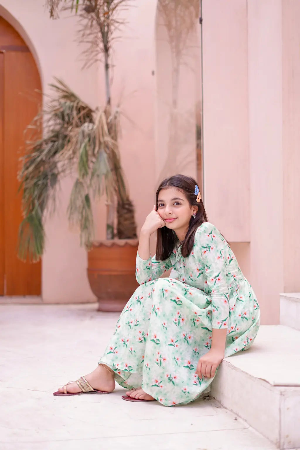 A young in a floral dress sitting on a white bench with a pink wall and plant in the background