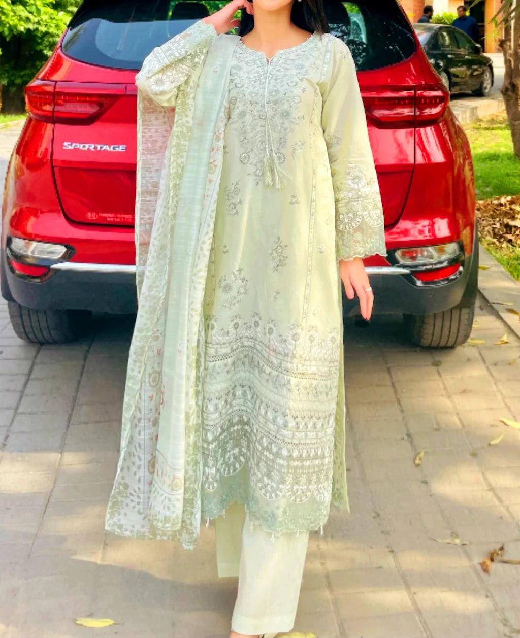 Person wearing a light green embroidered outfit standing in front of a red car.