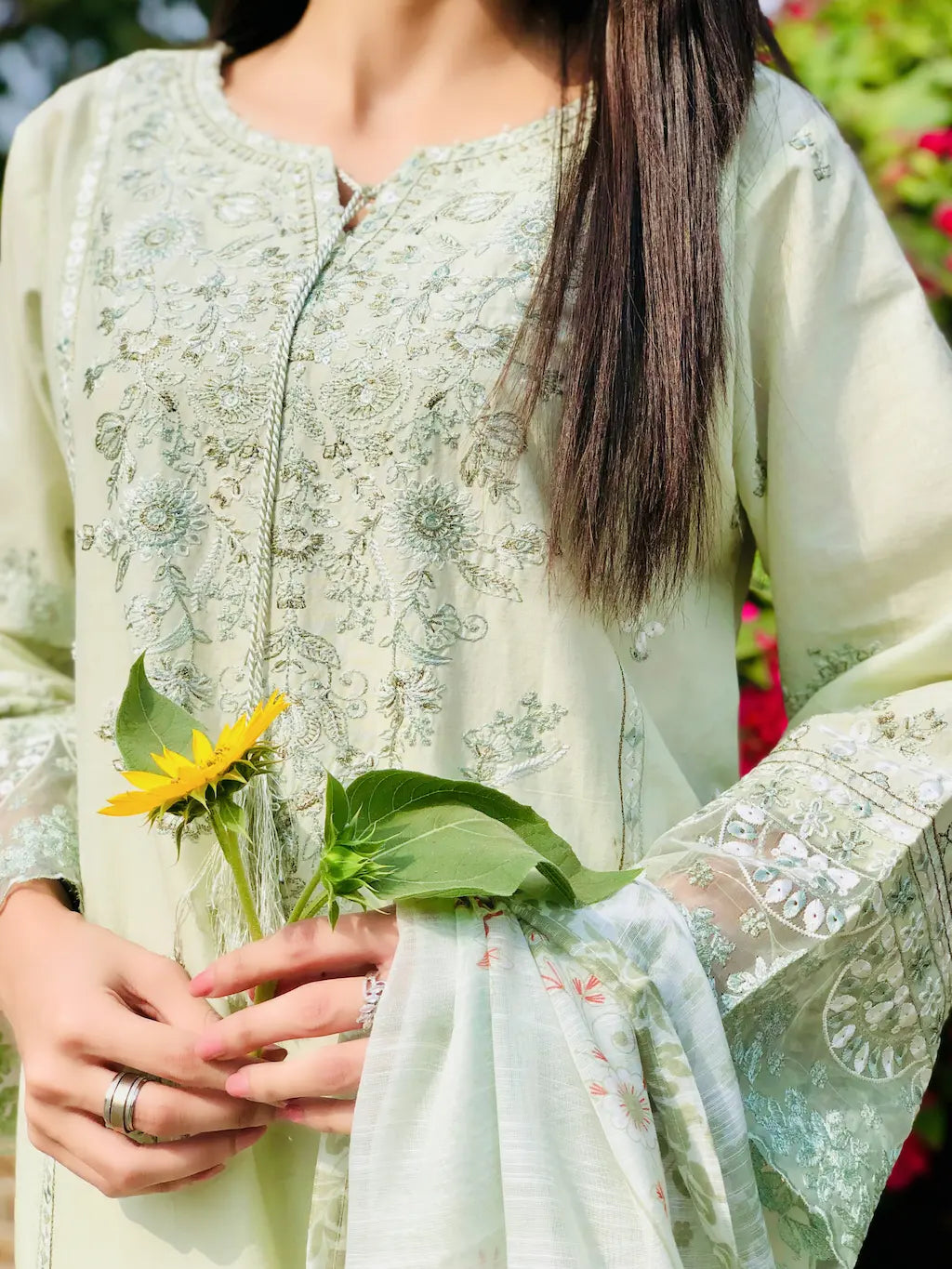 Person wearing a light green embroidered outfit holding a sunflower and leaves.