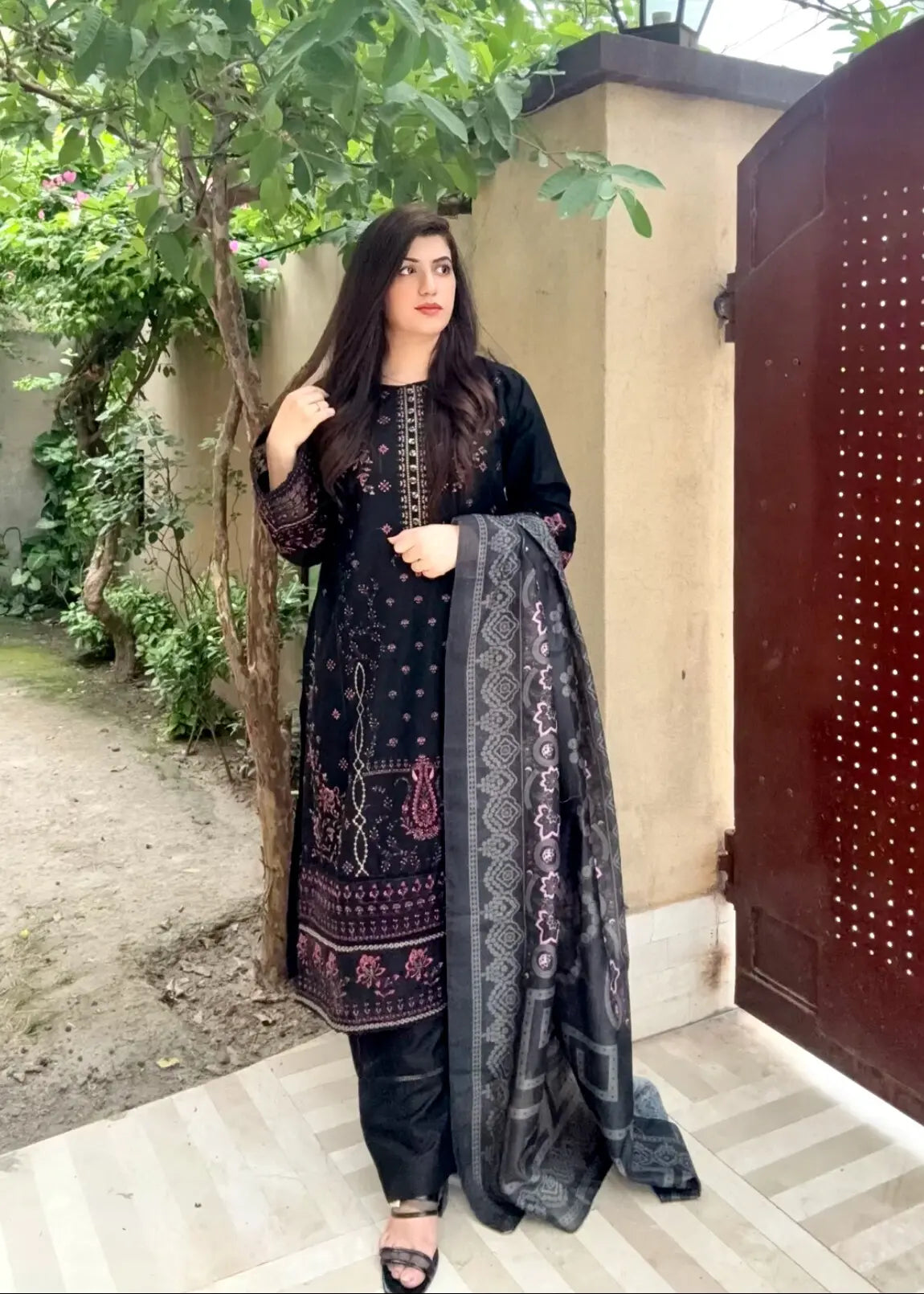 Woman in traditional black embroidered outfit with a dupatta, standing outdoors.