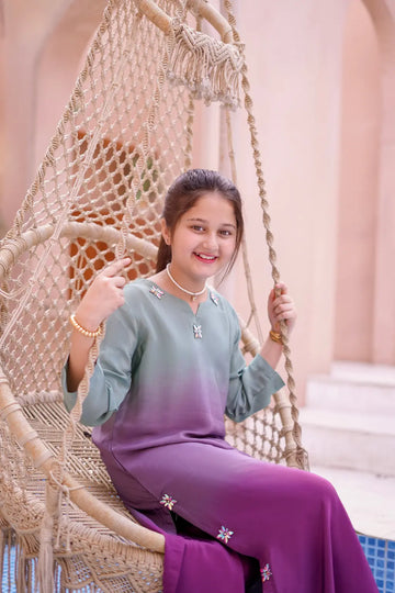 Girl sitting in a woven chair, wearing a green and purple outfit with floral details.