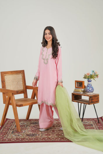 Woman in a pink traditional outfit with a green dupatta standing in a room with a wooden chair and small table.