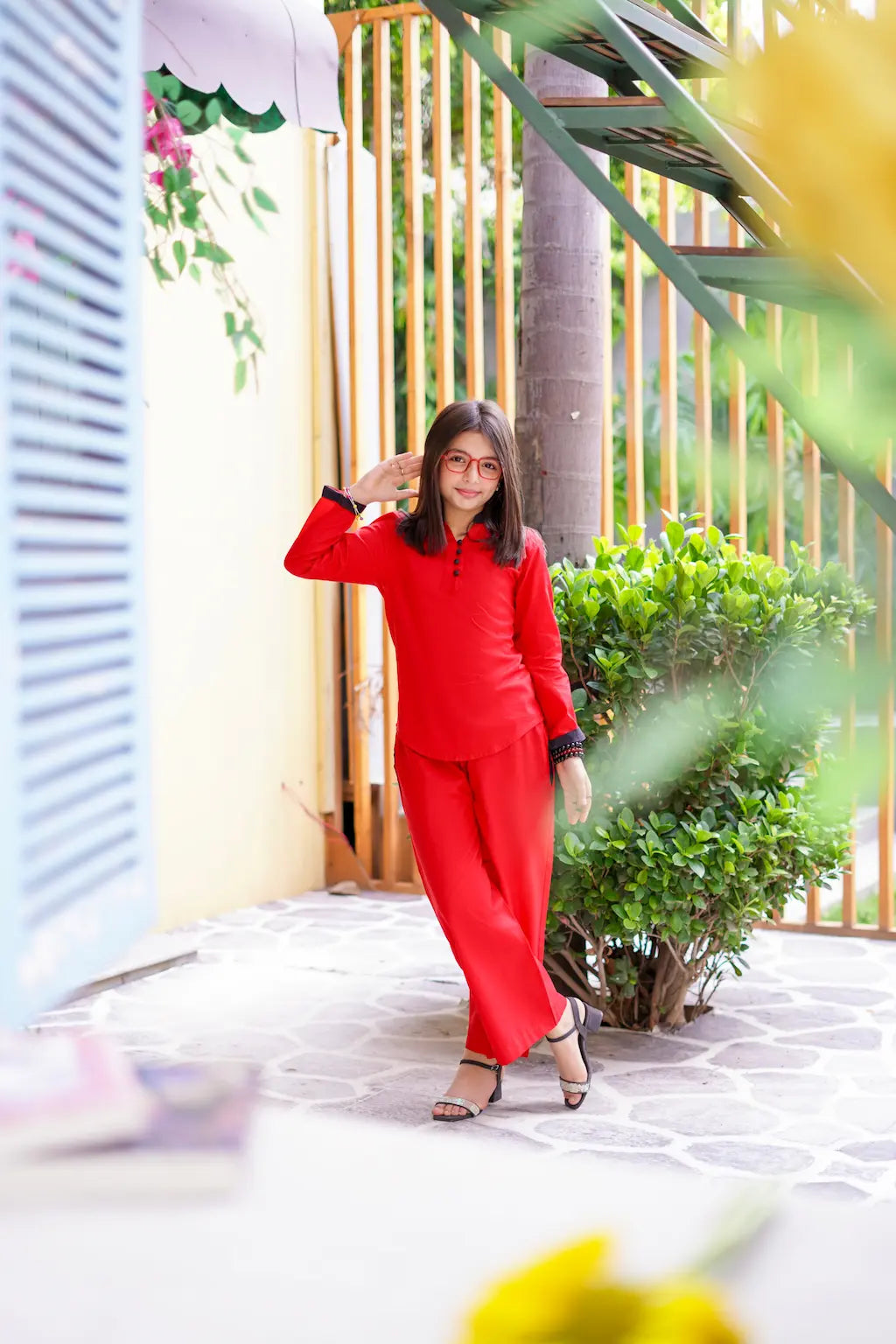 A young girl in a red outfit standing outdoors with plants and a colorful building in the background