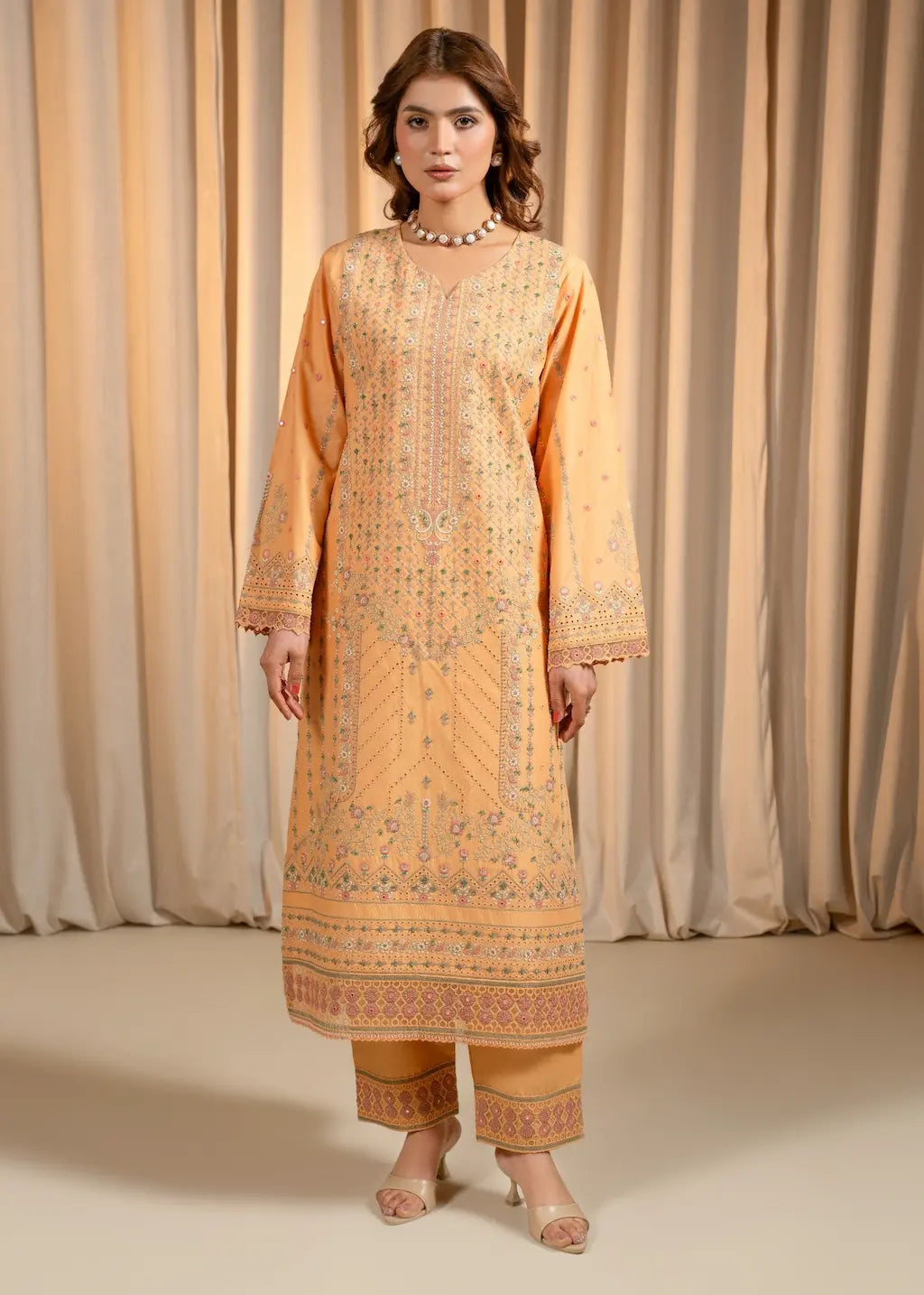 Woman wearing a peach-colored embroidered traditional outfit against a beige curtain background