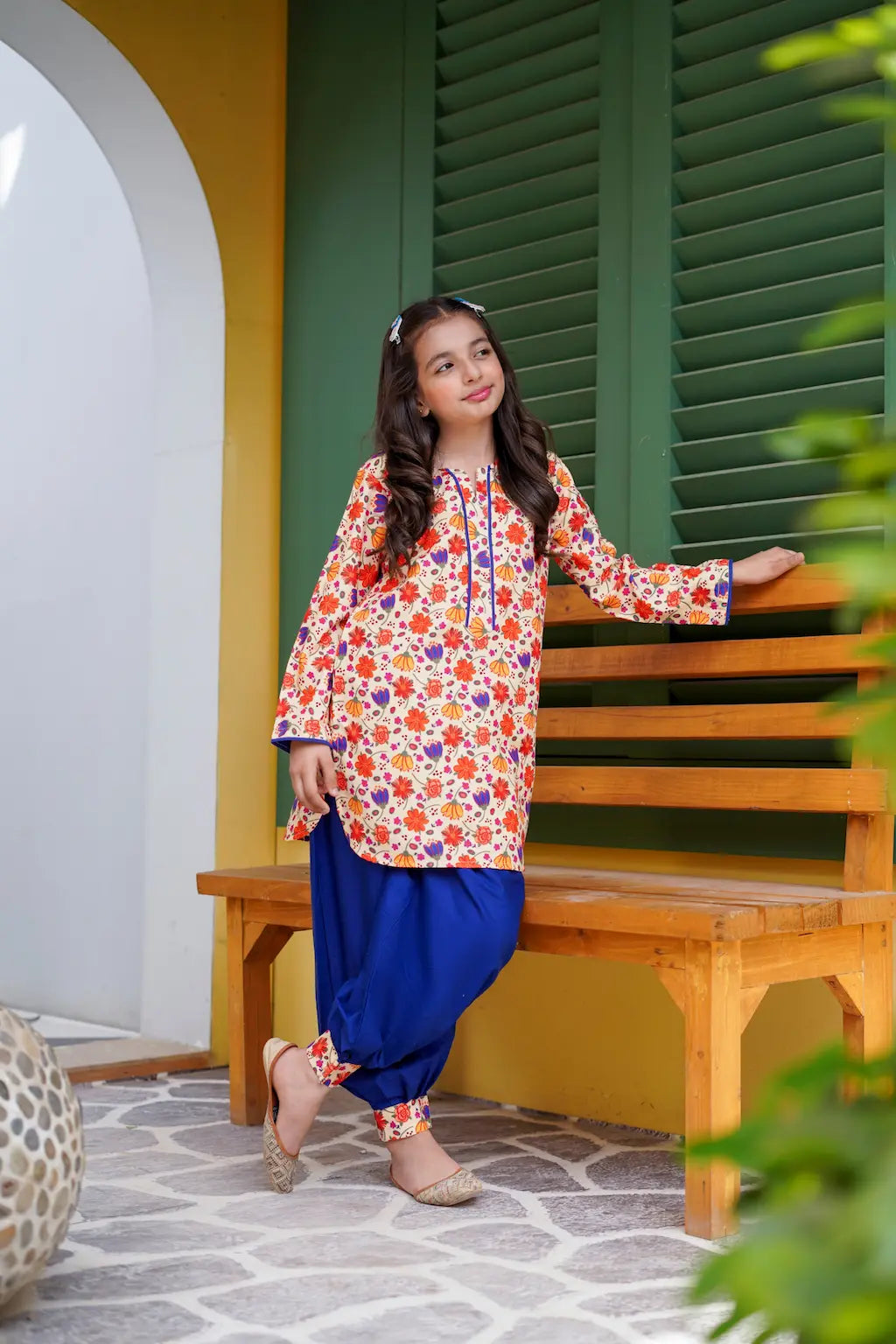 Young girl in a colorful floral top and blue shalwar sitting on a wooden bench.