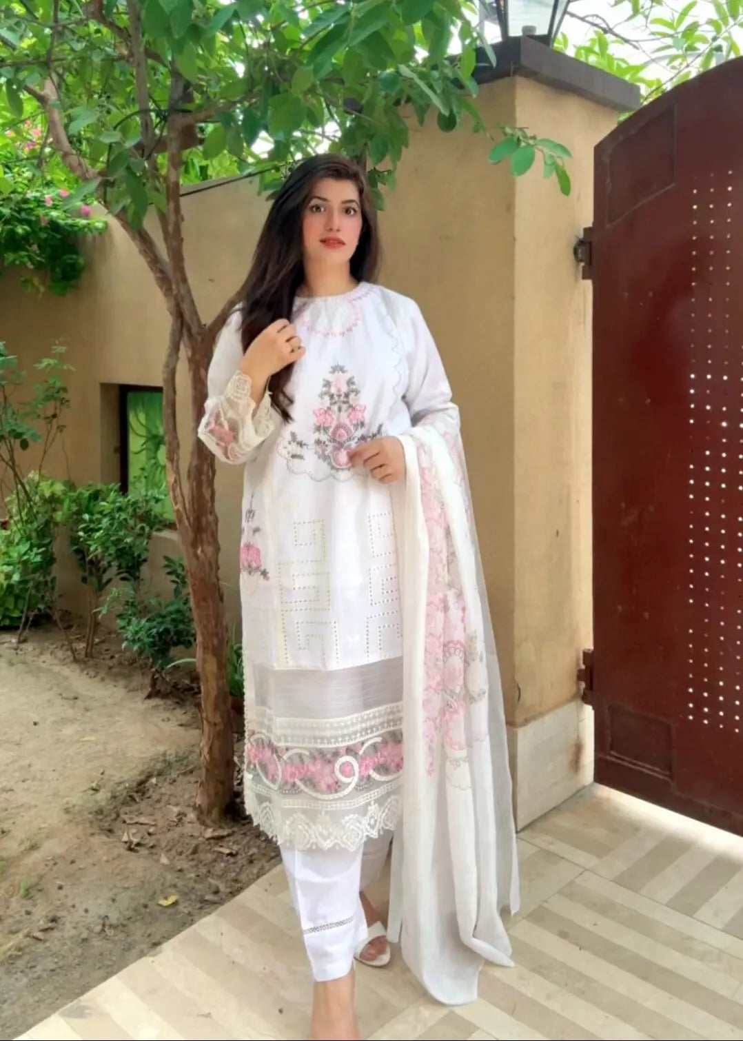 Woman in a white embroidered outfit standing outdoors near a tree and building.