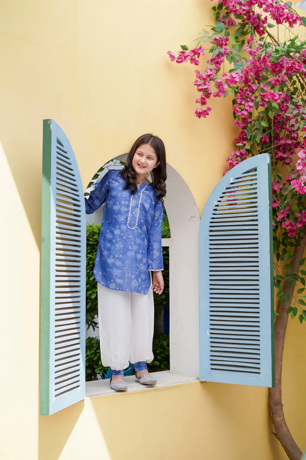 Young girl in a blue top and white pants standing in front of a yellow wall with a floral plant.
