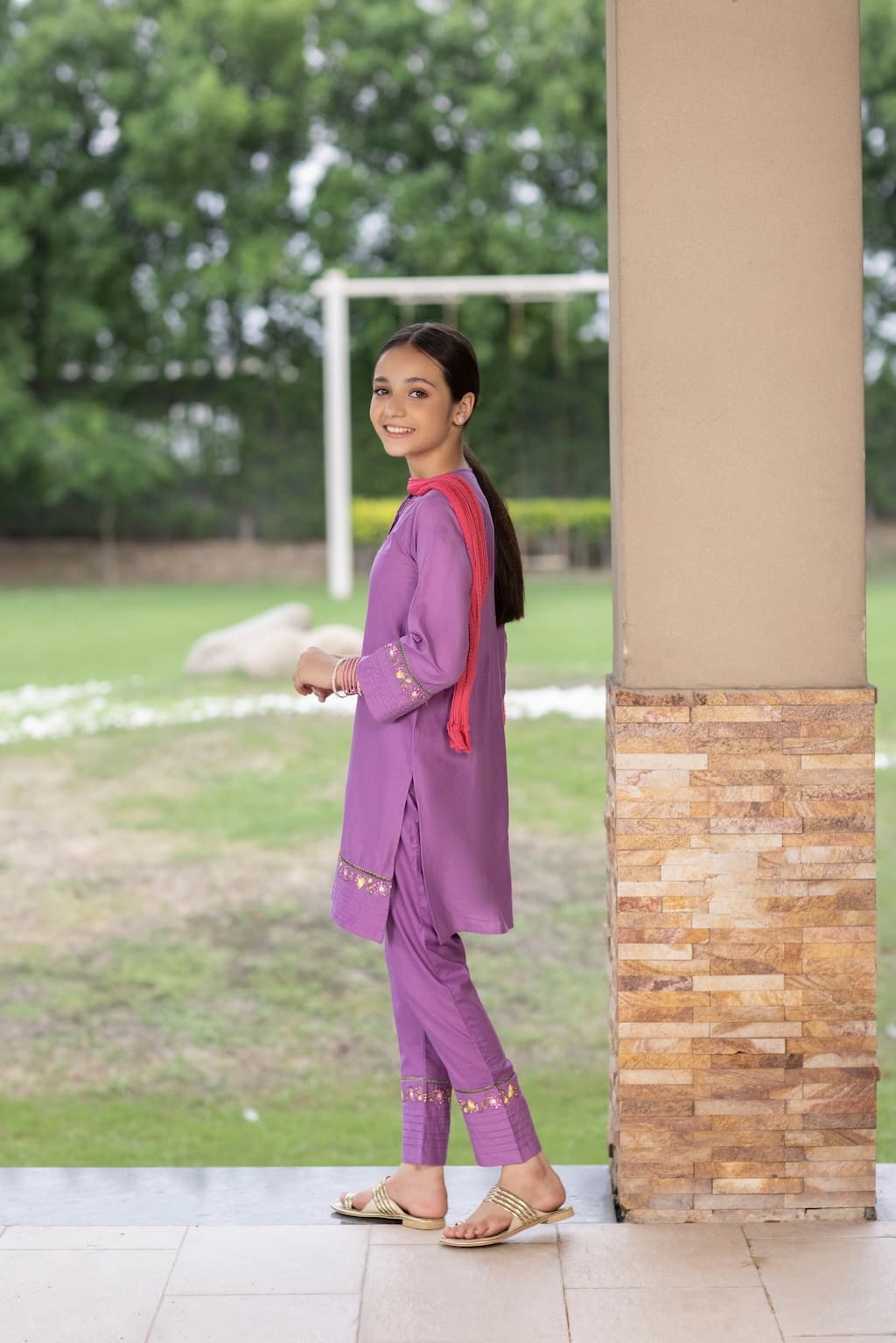 Girl in a purple outfit standing next to a stone column with greenery in the background