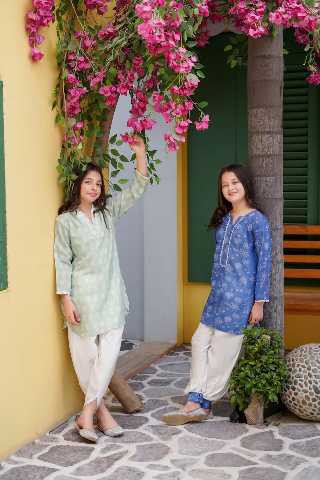 Two young girls in traditional outfits standing under a flowering archway.