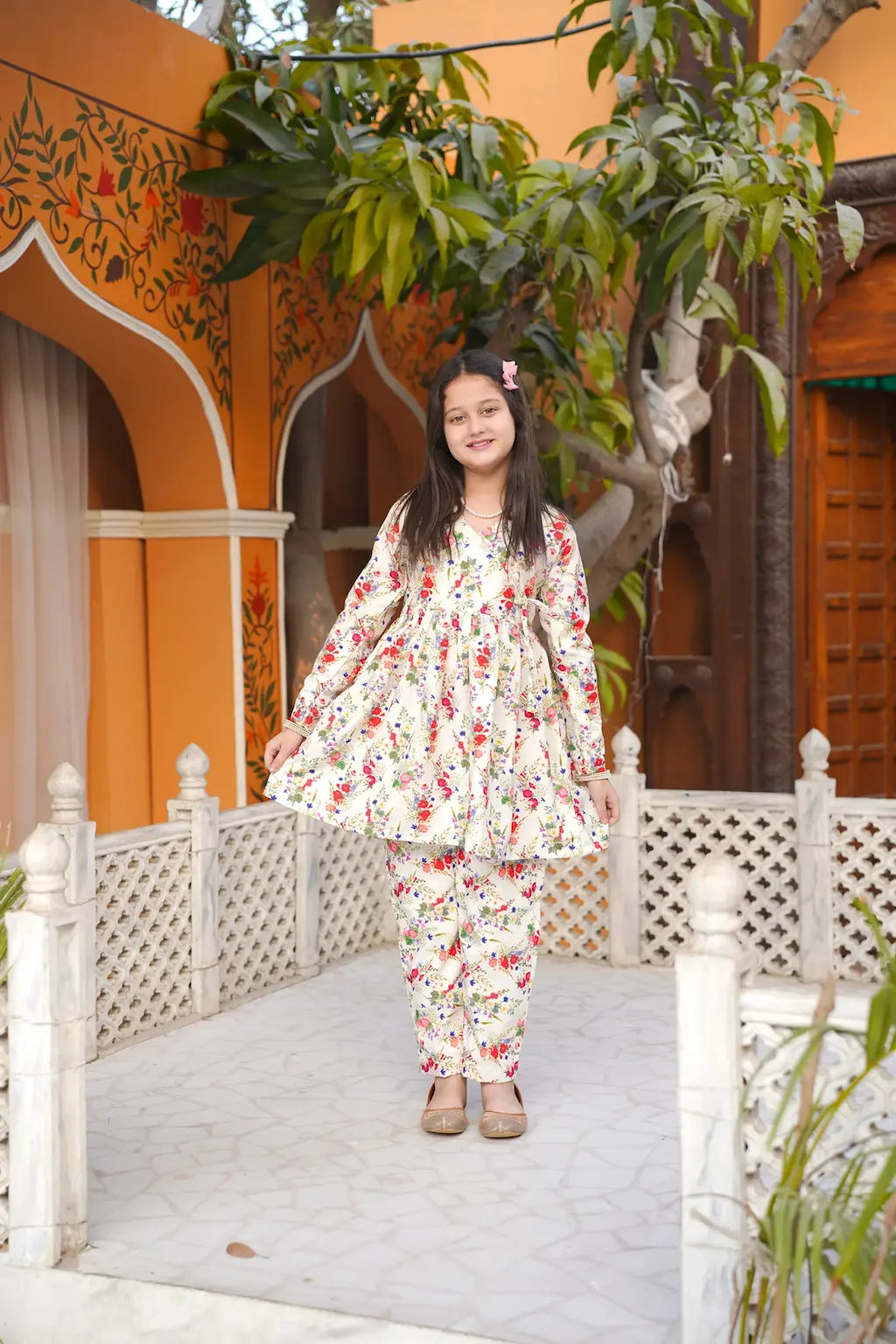 Young girl in a floral dress standing in front of an orange building with decorative elements.