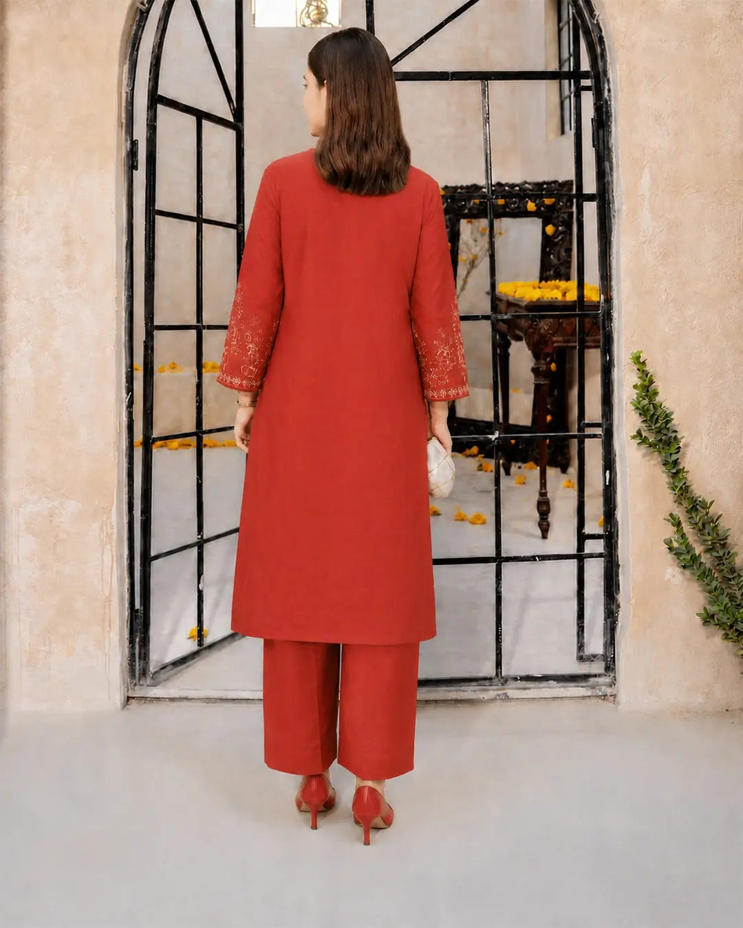 A girl viewed from behind wearing a bright red three-piece kurta set with matching wide-leg trousers and red heels, standing in front of a black metal arched doorway.