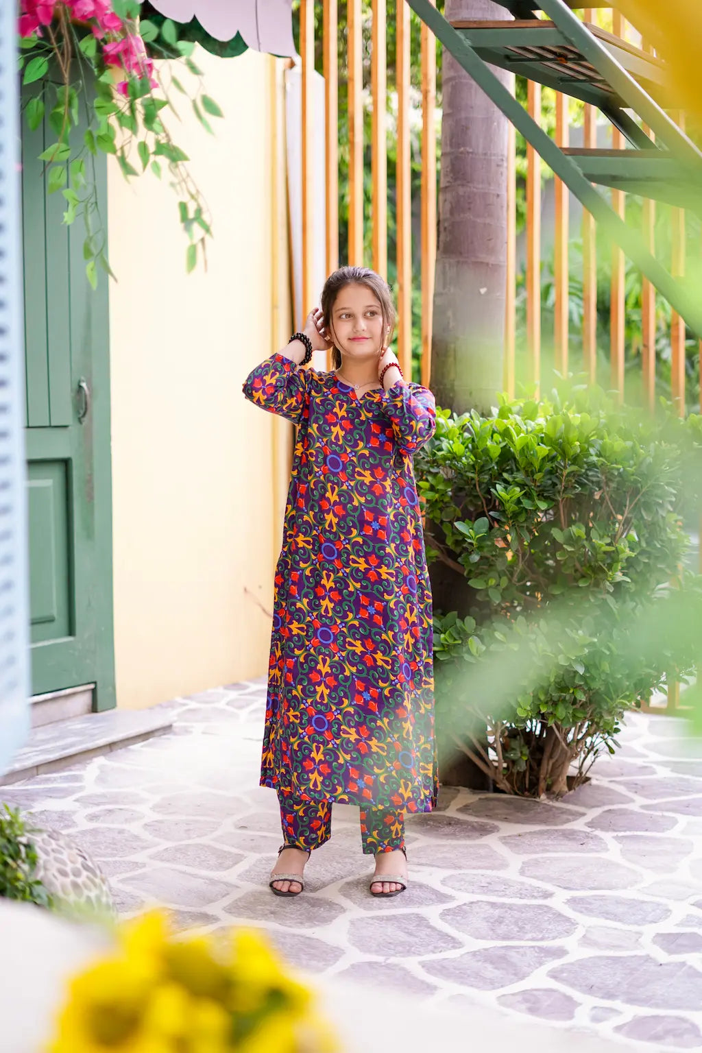 A girl in a colorful floral dress standing outdoors with greenery and a building in the background