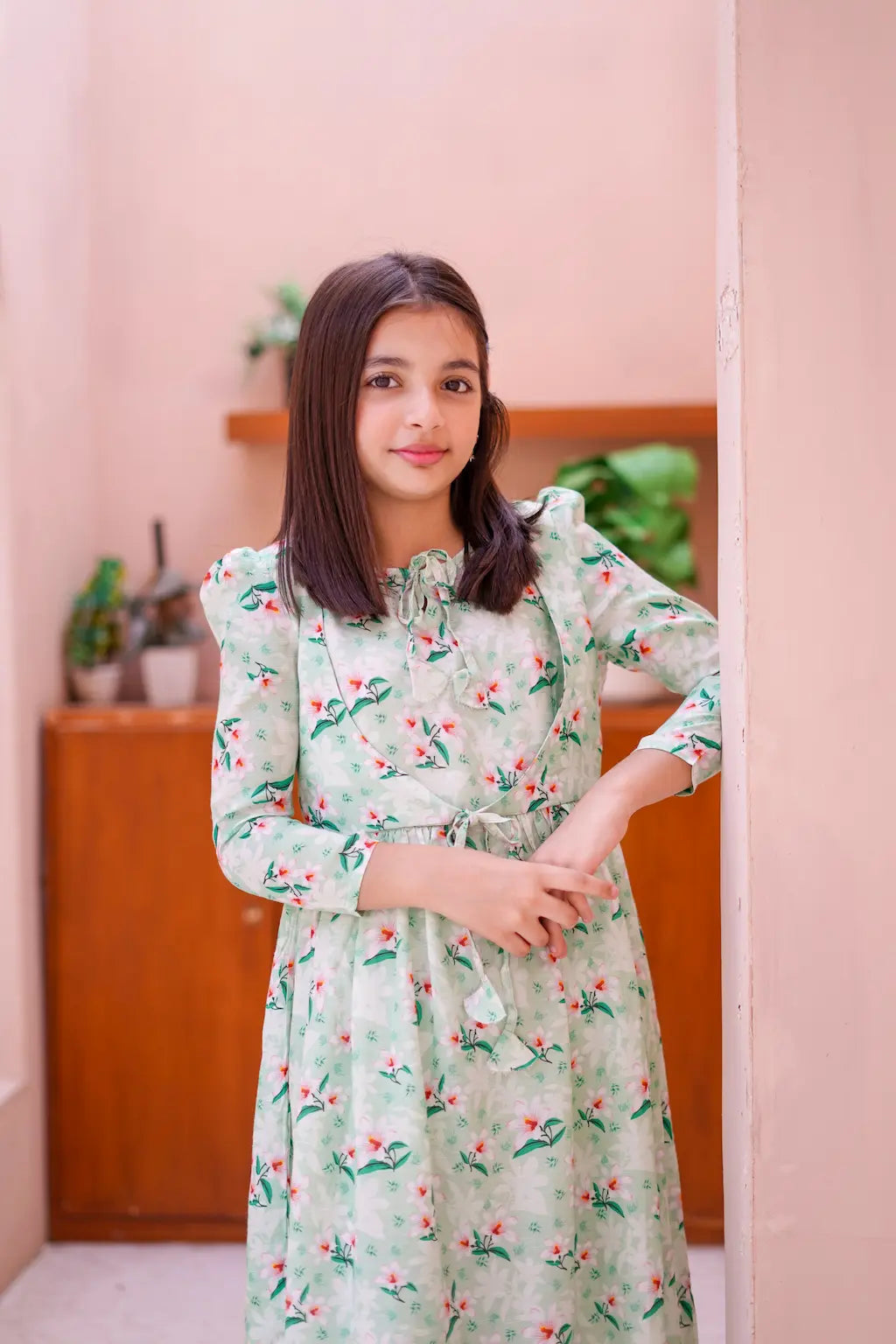 Young girl in a floral dress standing in a room with a pink wall and wooden cabinet.