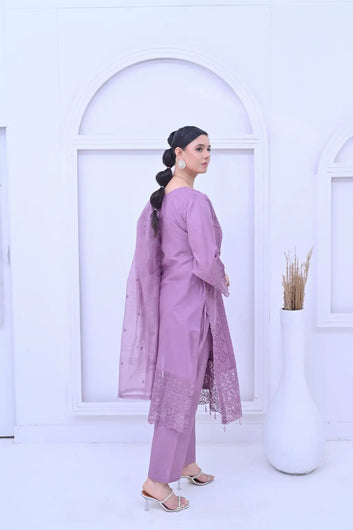 A side-profile view of a woman wearing a lavender-colored three-piece embroidered suit. The outfit includes a kameez with an intricate lace border and cutwork sleeves, matching straight trousers, and a sheer embroidered dupatta draped over the shoulder. She is posed against a white minimalist background.