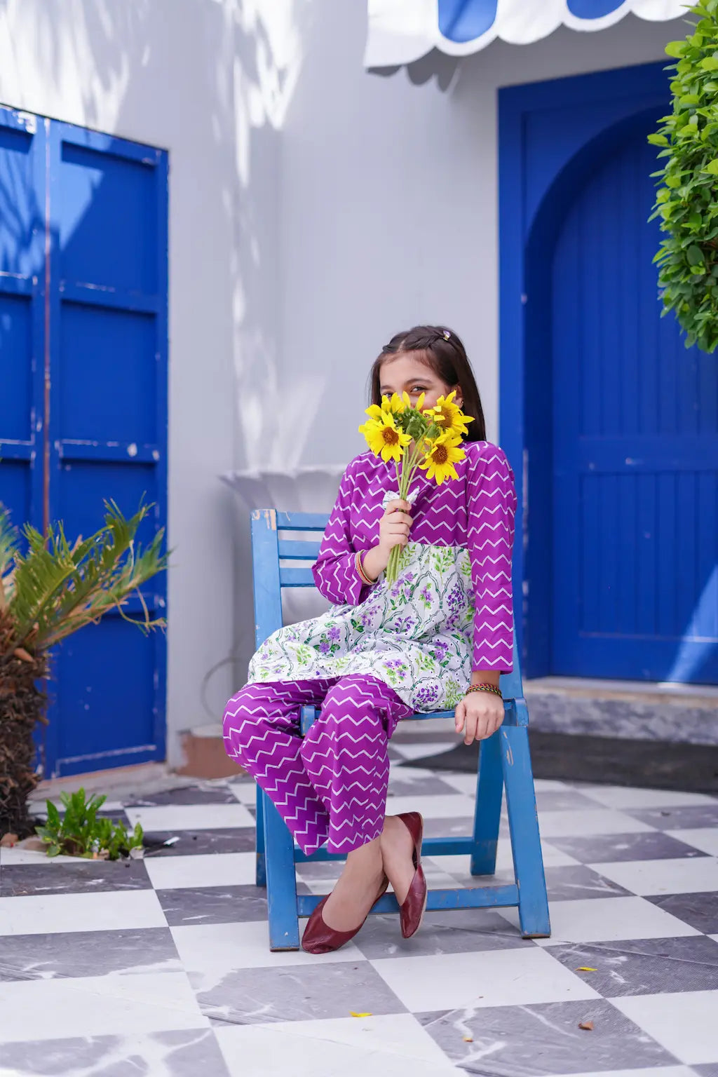 A young girl holding sunflowers sitting on a blue chair in front of a white wall with blue doors.