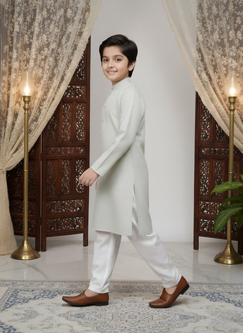 Young boy in a white kurta standing in a decorated room with curtains and lamps.