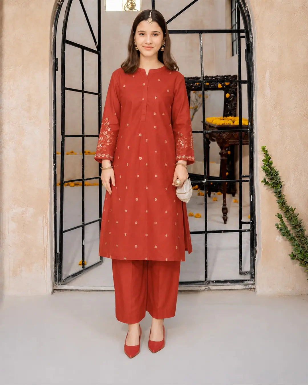 A girl modeling a rust-red kurta and trouser set with small gold motifs, standing in front of a black metal arched gate on a beige wall.