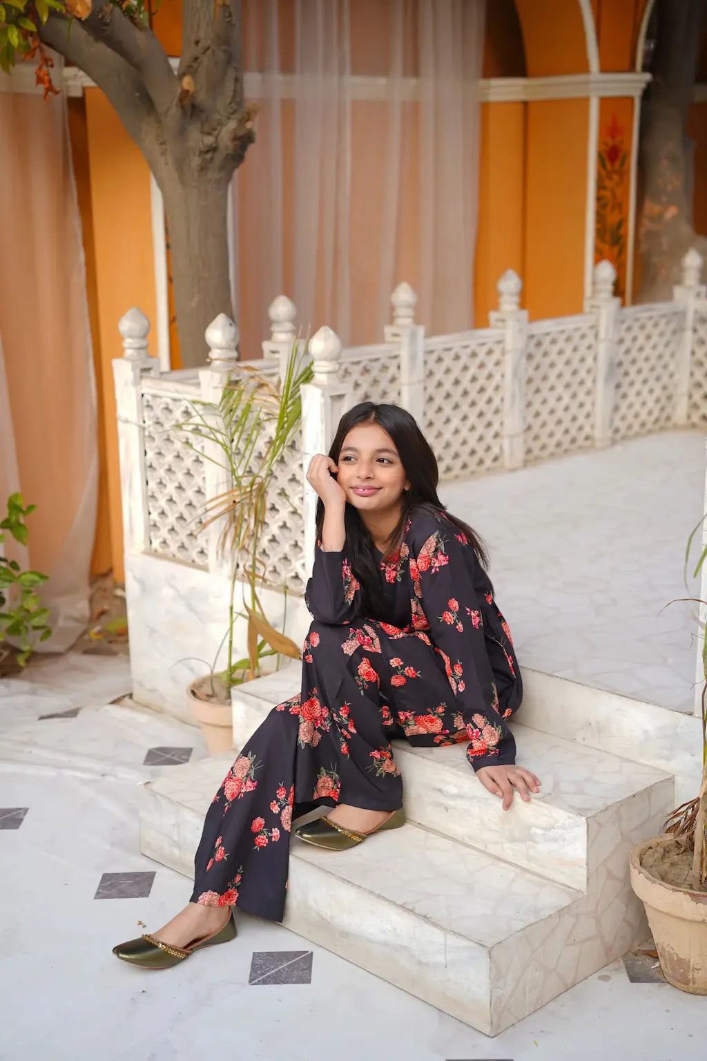 Young girl in a floral dress sitting on stone steps with an orange building in the background