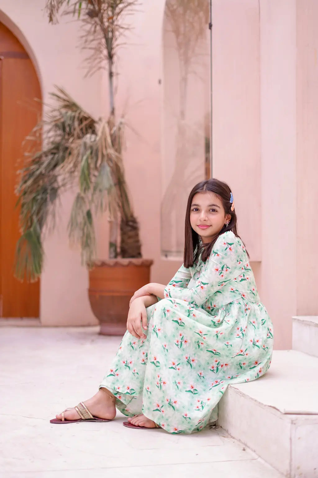 Young girl in a floral dress sitting on a white couch with a pink wall and plant in the background