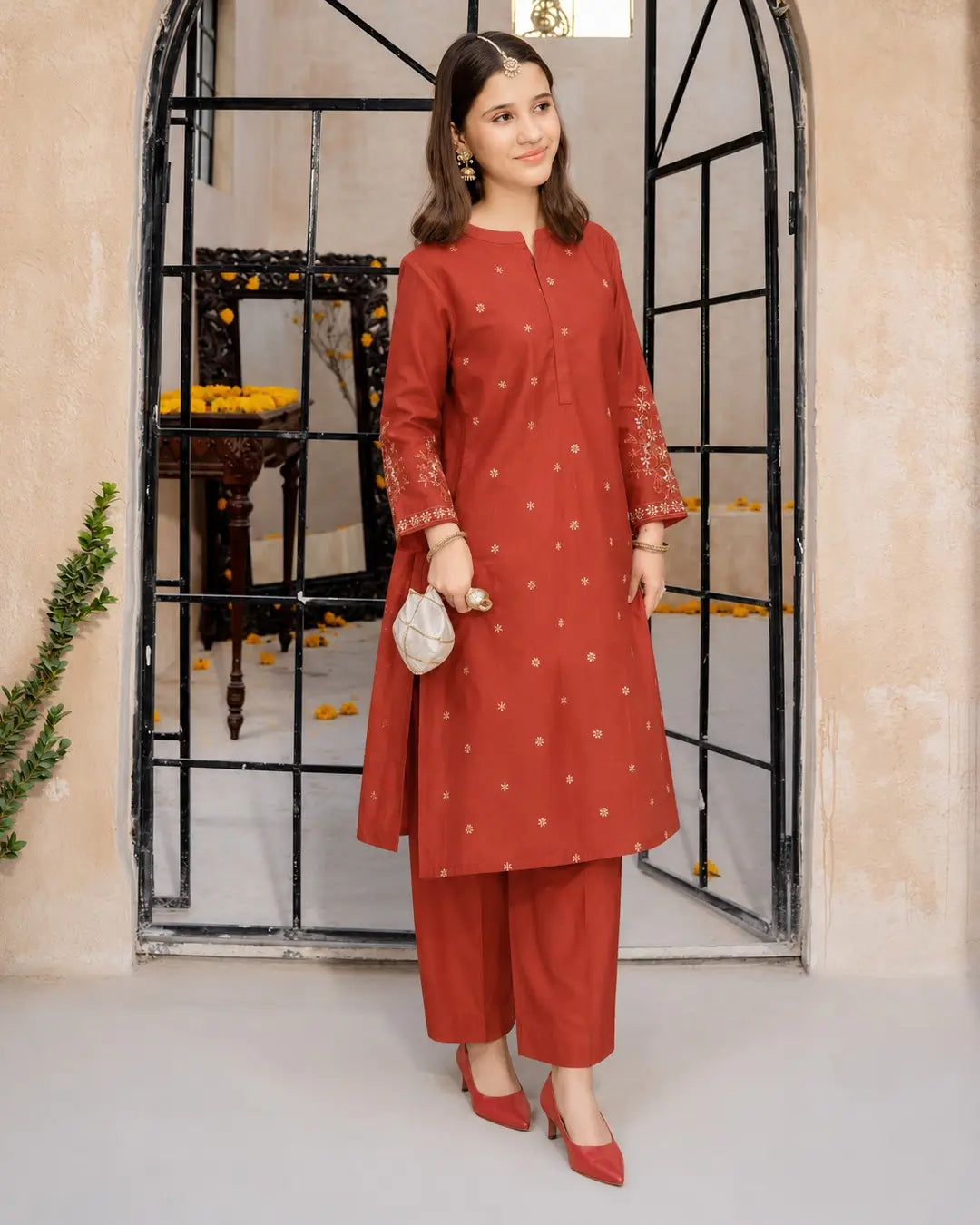 A girl models a rust-red three-piece kurta set with matching pants and red pointed-toe heels, holding a small white clutch bag, standing in front of a black metal arched door.
