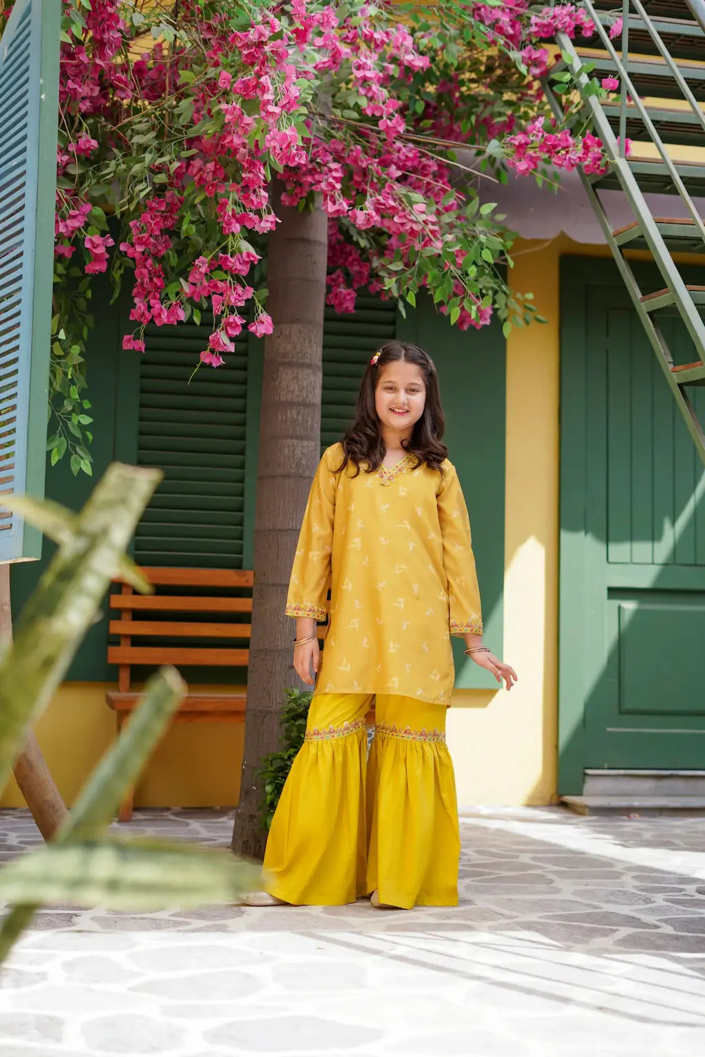 A young girl in a yellow traditional outfit standing under a flowering tree with a green and yellow building in the background.