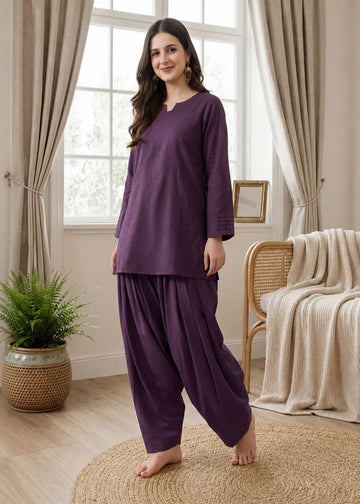 Woman wearing a purple outfit standing in a bright room with curtains and a plant.
