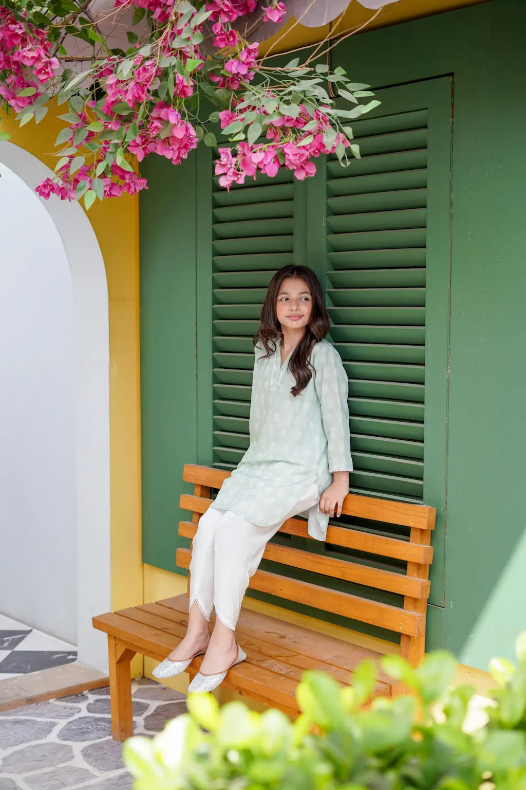 A young girl sitting on a wooden bench in front of a green shuttered door with pink flowers above.