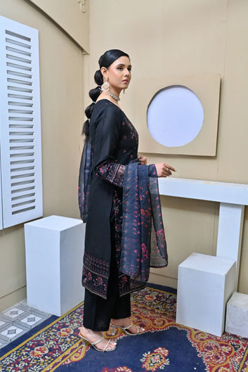 Model in a black three-piece embroidered suit with pink floral details, lace-trimmed sleeves, and a printed dupatta, posing in a minimalist studio setting.