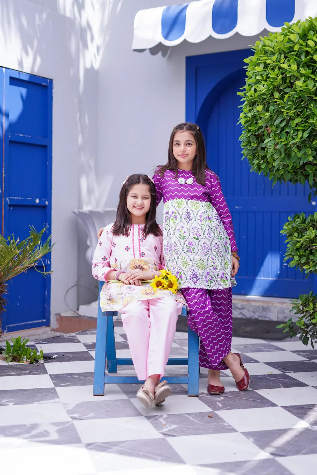 Two young girls in matching outfits standing in front of a blue door with greenery.