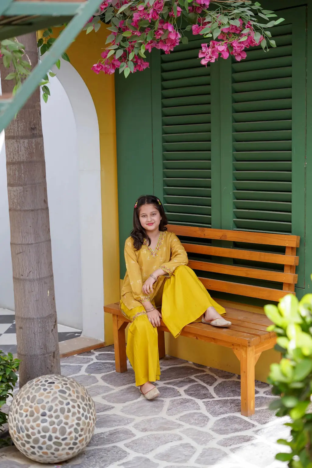 A young girl in yellow outfit sitting on a wooden bench under a colorful building with pink flowers.