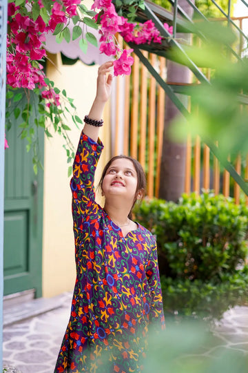 Young girl in a colorful dress reaching up to touch pink flowers outdoors.
