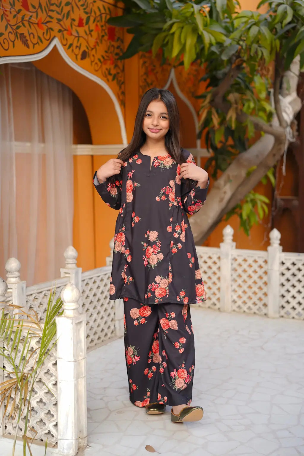 Young girl in a floral outfit standing in front of an orange building with white lattice work.