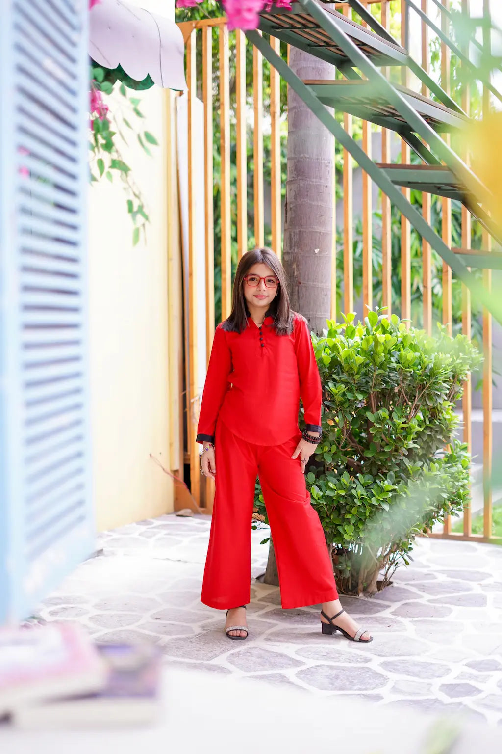 A young girl in a red outfit standing outdoors with plants and a colorful structure in the background