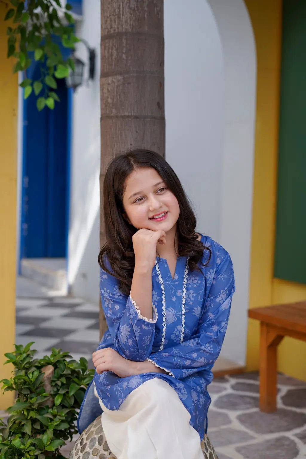 Young girl in a blue traditional outfit sitting outdoors with a colorful background