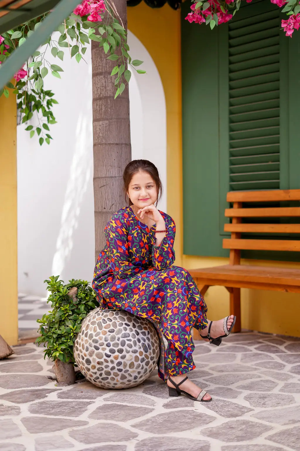A young girl in a colorful outfit sitting on a decorative ball in a garden setting.
