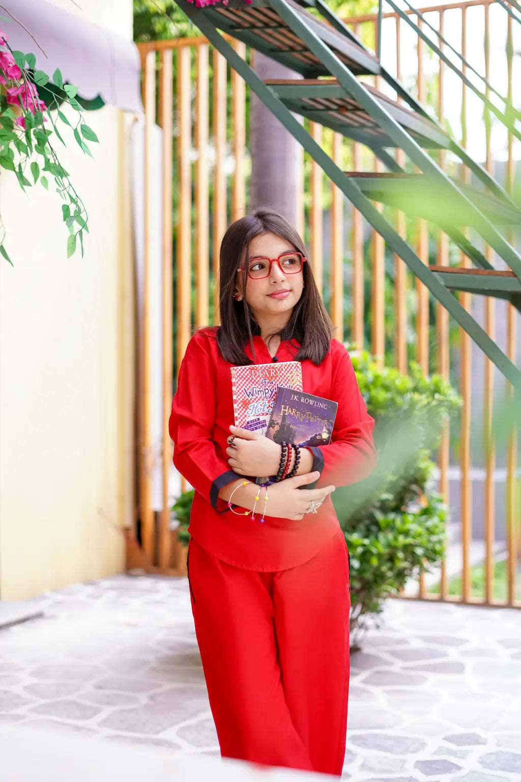 A young girl in a red outfit holding books in an outdoor setting with plants and a staircase.