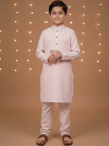Young boy wearing a light pink kurta with white pants against a warm-toned background with string lights.