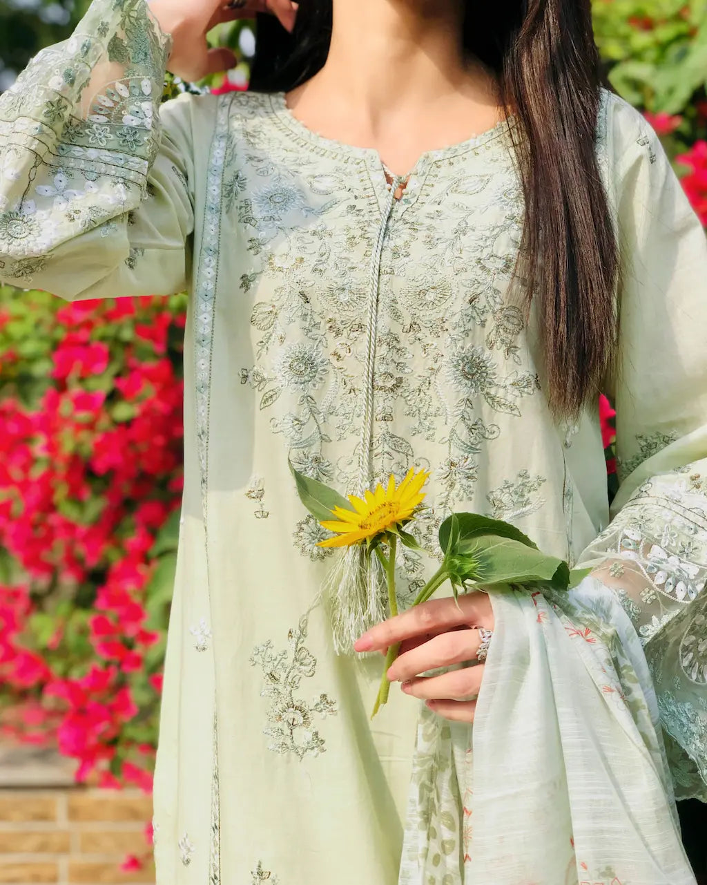 Woman in a light green embroidered outfit holding a yellow flower with a blurred floral background