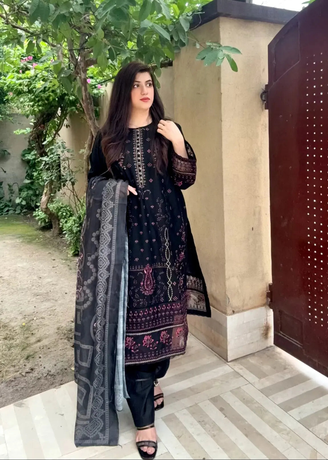 Woman in traditional black outfit standing outdoors with greenery in the background