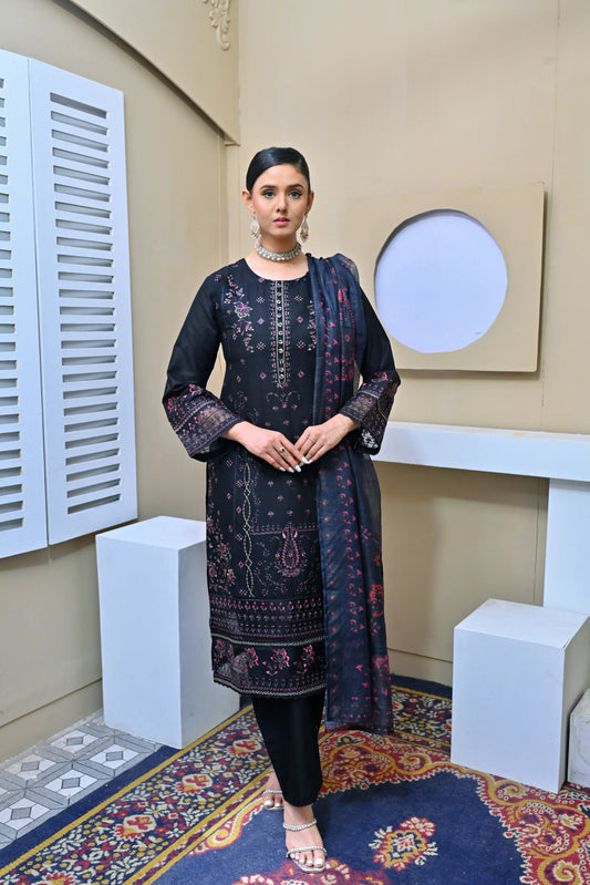 Model in a black three-piece embroidered suit with pink floral details, lace-trimmed sleeves, and a printed dupatta, posing in a minimalist studio setting.