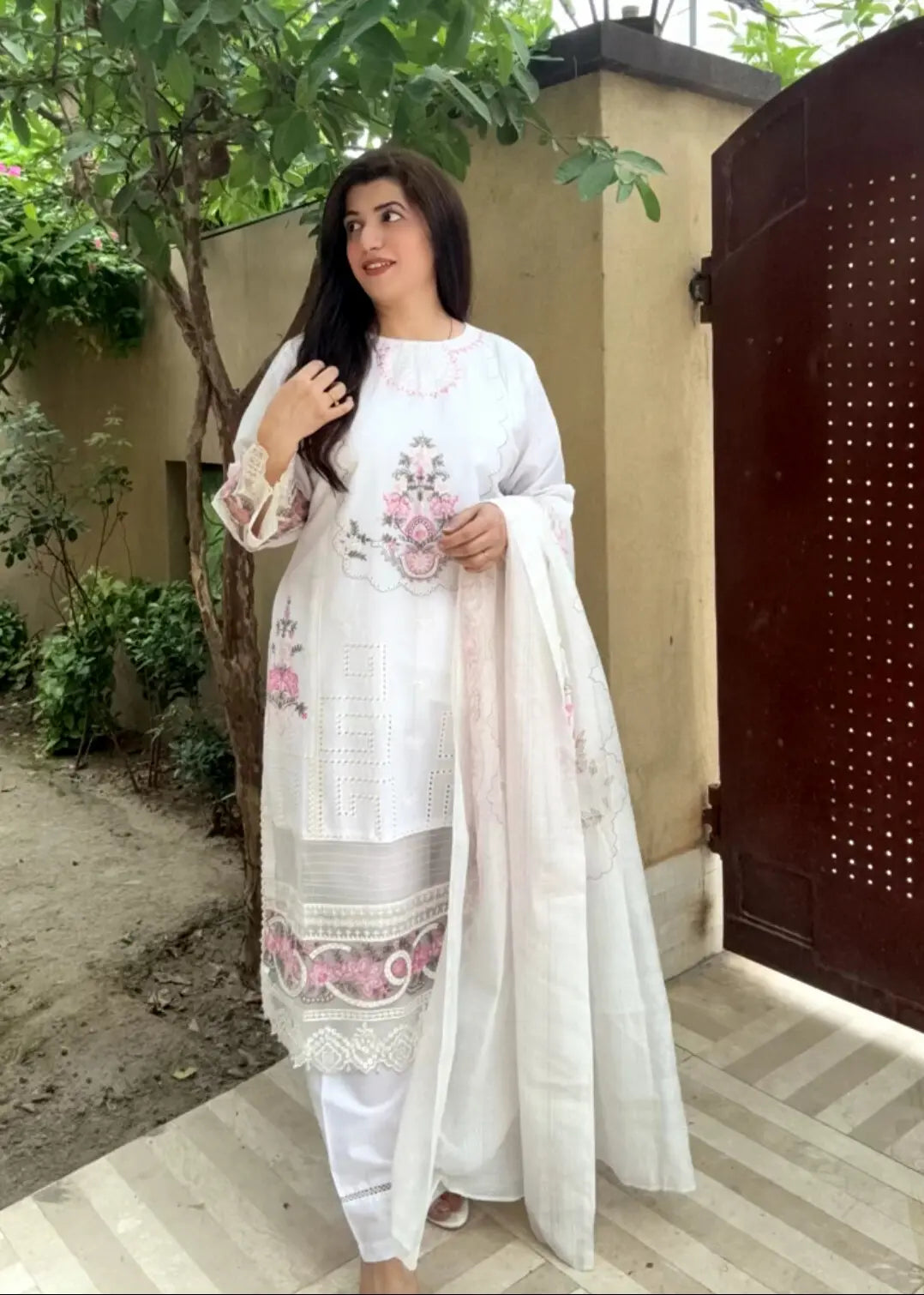 Woman in a white embroidered outfit standing outdoors with greenery and a gate in the background